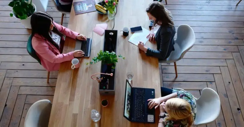 A view from above a work table in the Stratice office. Three women sit at this rectangular table, working on laptops and paperwork.