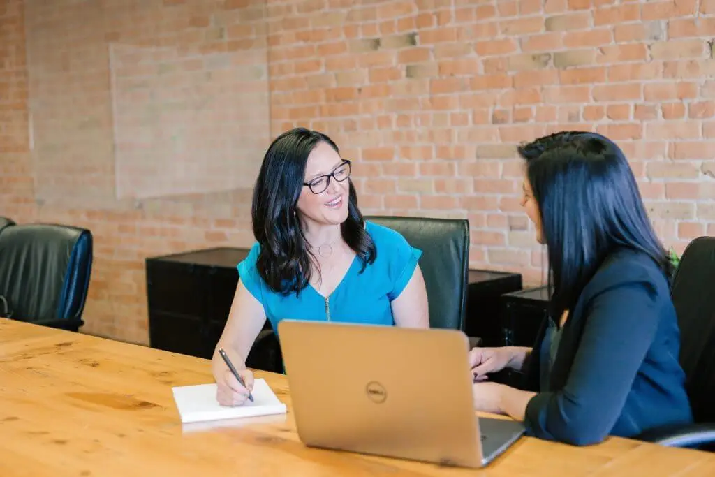 Two women sitting at a table, talking and taking notes.