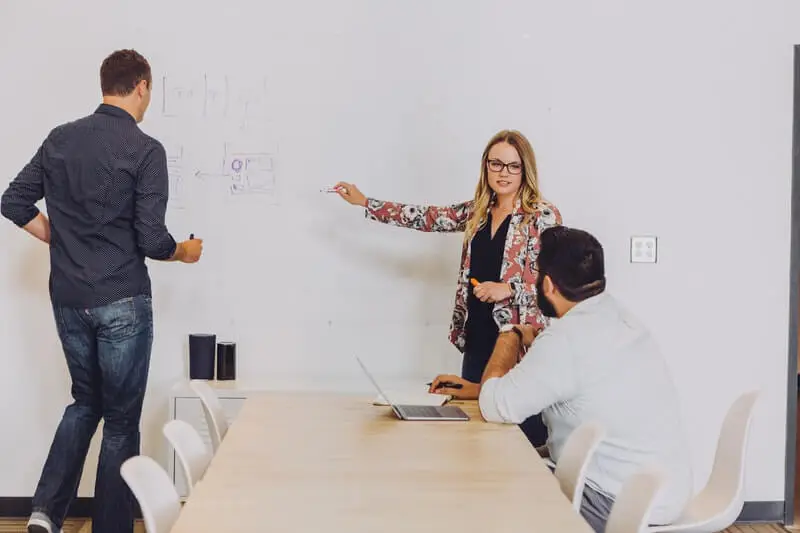 Two coworkers standing at a whiteboard, drawing and talking to their other teammate who is sitting at a table.