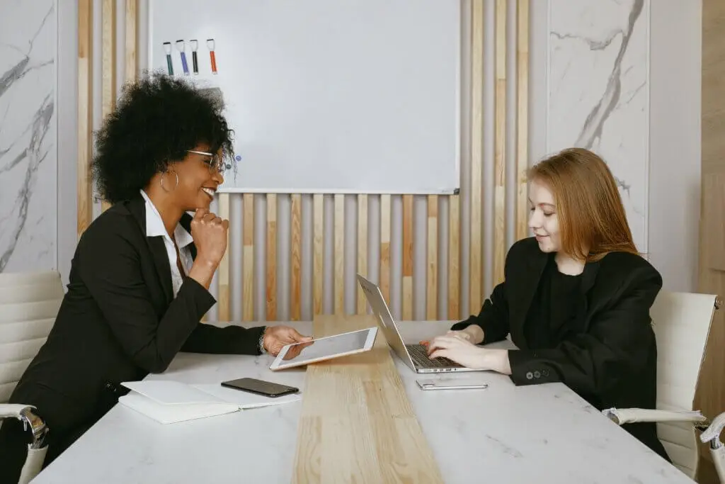 Two young women sit at a table across from each other, one on her tablet and one on her laptop.