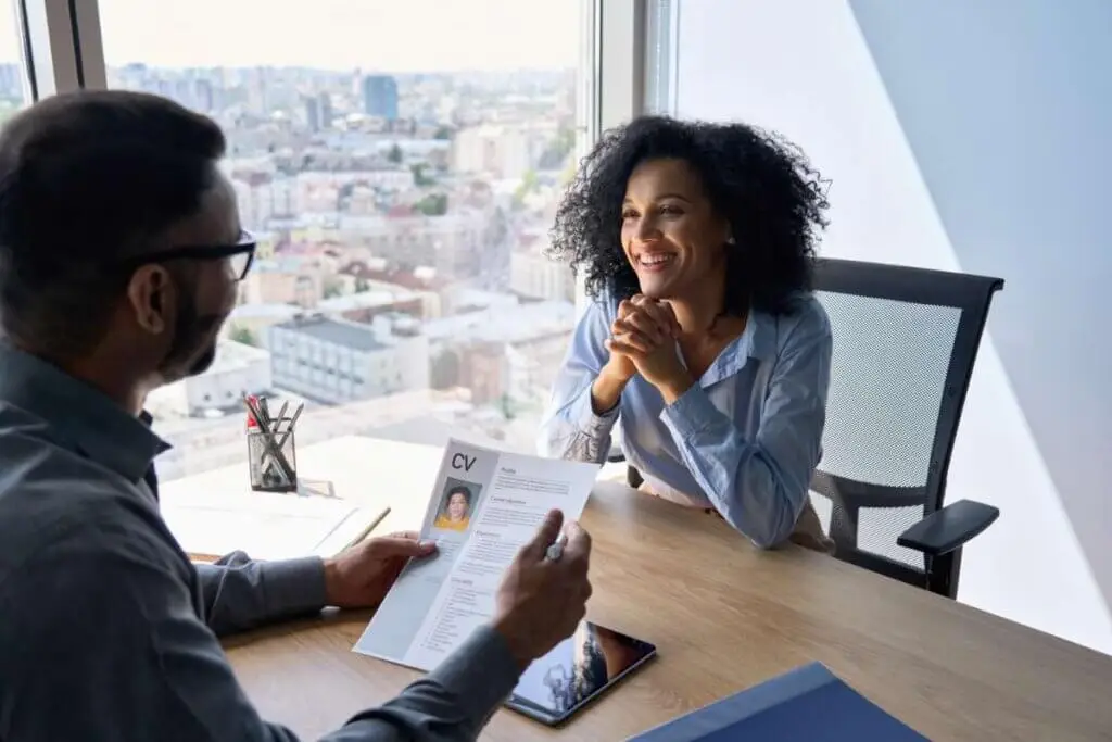 A woman sits at a table in a high-rise office in an interview. The interviewer is reviewing her CV.